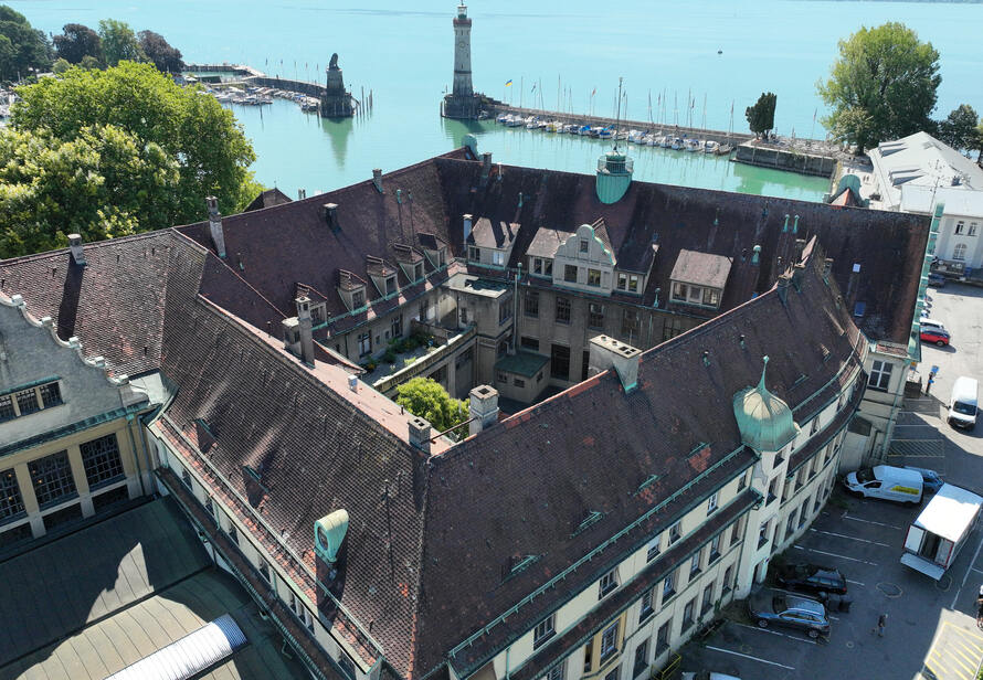 Die Drohnenaufnahme zeigt den Bahnhof Lindau mit seinen komplexen Dachflächen direkt am Bodensee. Im Hintergrund sind der Hafen Lindau, der Leuchtturm und der Bayerische Löwe zu sehen.
